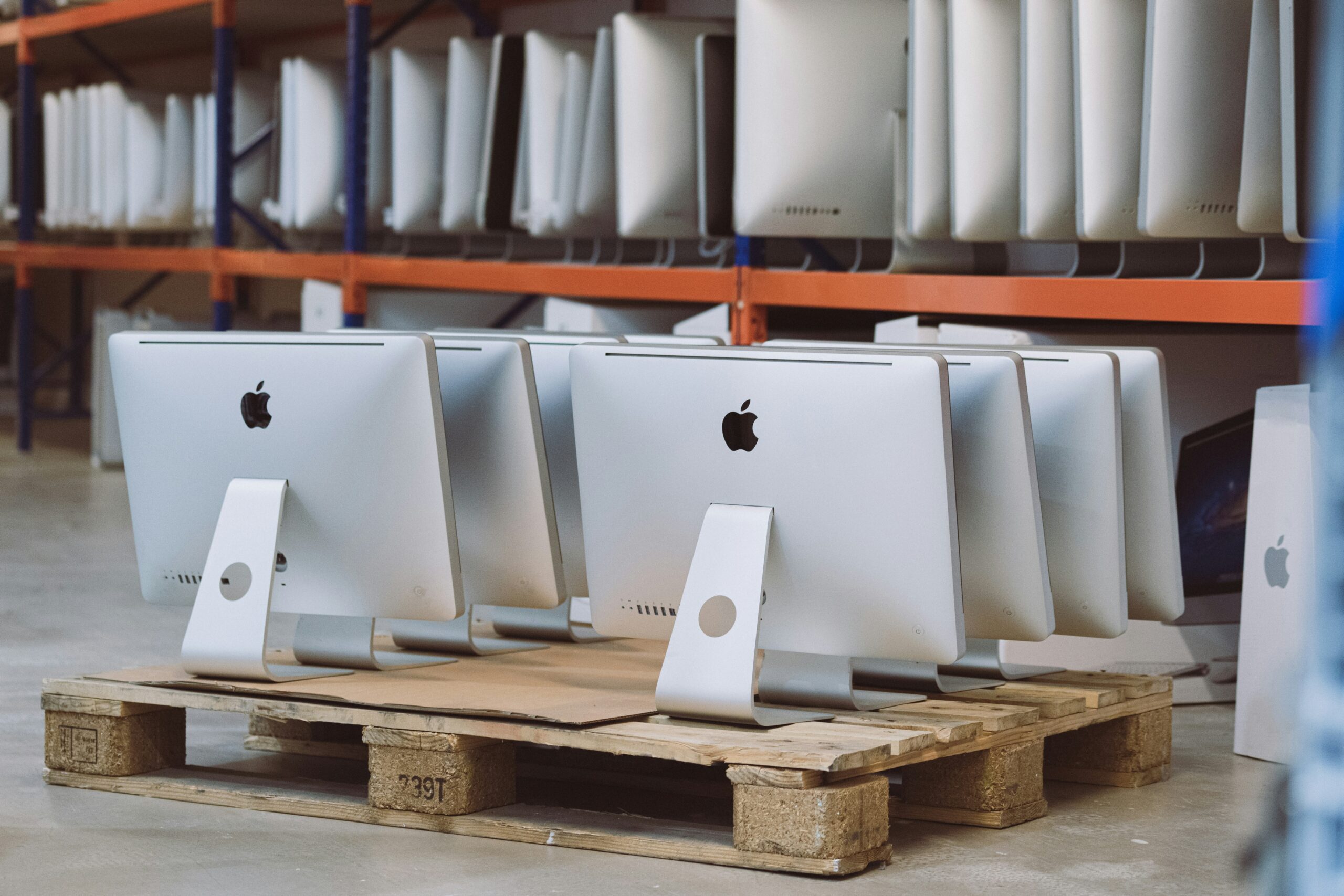 Rows of Apple iMac computers stacked on wooden pallets in a warehouse or storage area.