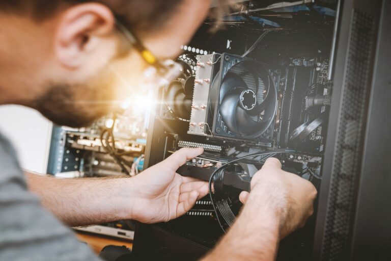 A bearded man wearing glasses leans over an open desktop PC case, using both hands to connect a cable to a component. The interior shows a large CPU cooler with heat pipes and fans, visible RAM slots, and cable routing, with a warm lens flare in the background suggesting a workshop setting.