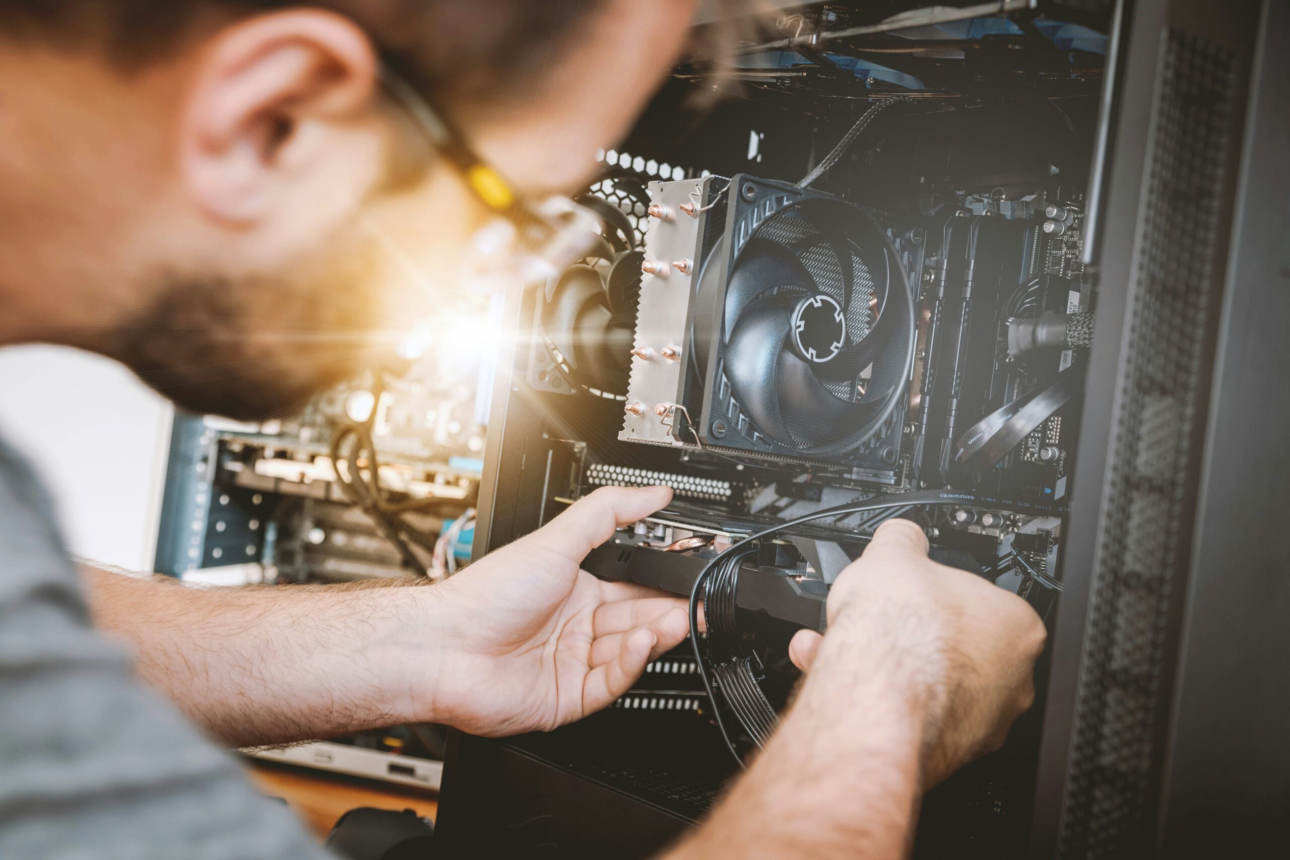 A bearded man wearing glasses leans over an open desktop PC case, using both hands to connect a cable to a component. The interior shows a large CPU cooler with heat pipes and fans, visible RAM slots, and cable routing, with a warm lens flare in the background suggesting a workshop setting.